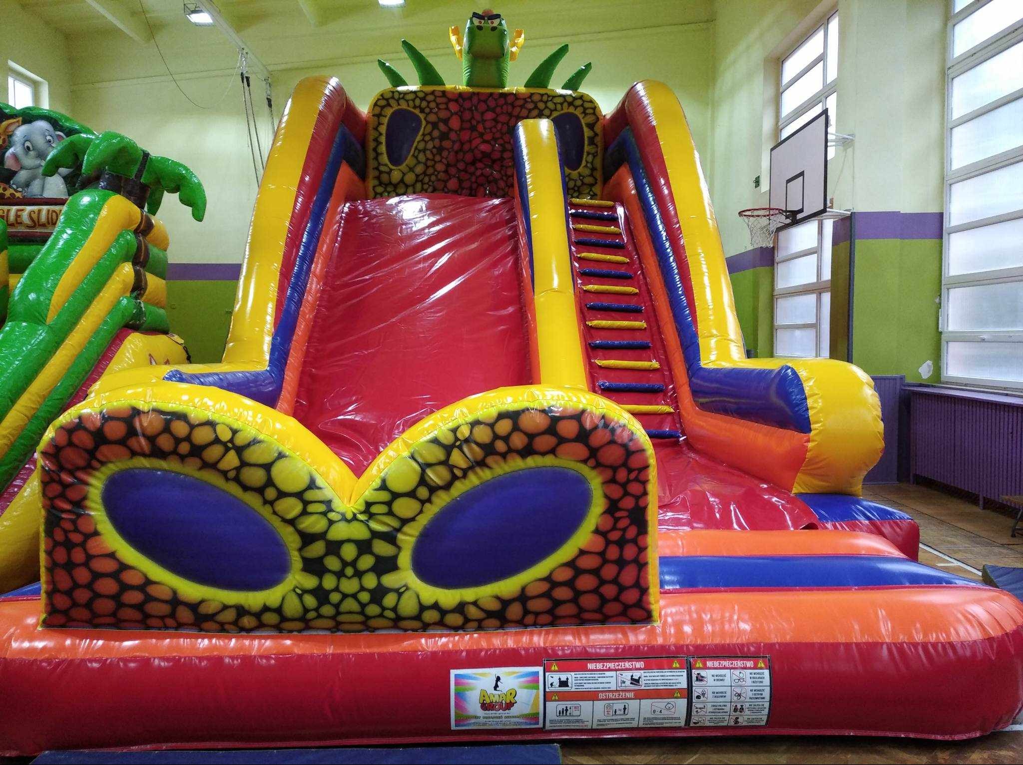 Kolorowa, dmuchana zjeżdżalnia z motywem dinozaura w sali gimnastycznej. Widoczne schody, zjeżdżalnia i elementy dekoracyjne. Znak ostrzegawczy na dole.
