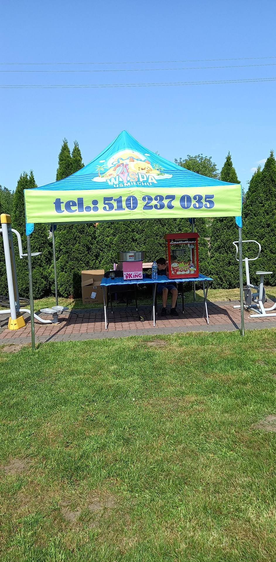 Stoisko z automatem do popcornu pod turkusowym namiotem z numerem telefonu, ustawione na trawie obok siłowni plenerowej w słoneczny dzień.