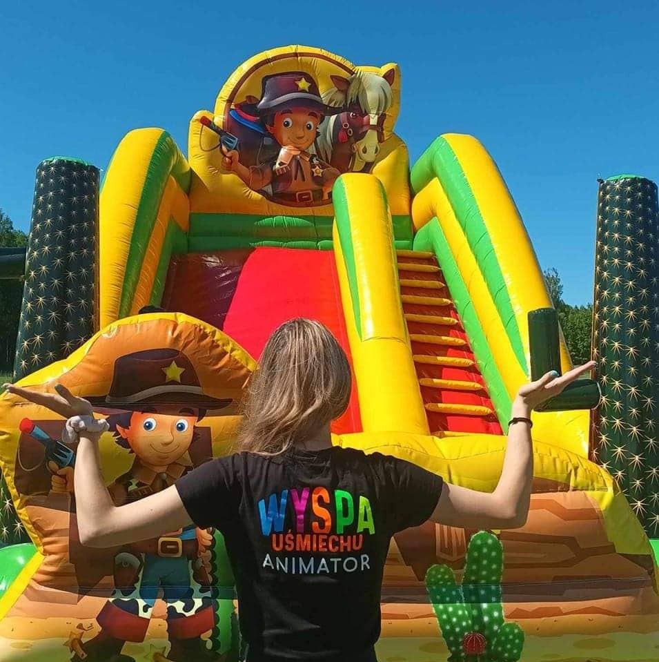 Kolorowy dmuchany zamek w stylu western z postacią kowboja, prezentowany przez osobę w koszulce z napisem 'Animator'. Słoneczny dzień, błękitne niebo.