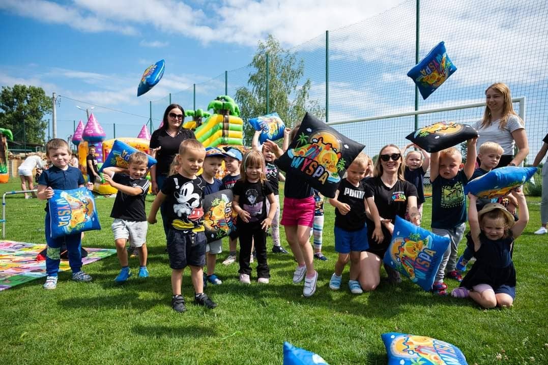 Grupa dzieci na trawie trzymająca poduszki z logo 'Wyspa', w tle dmuchany zamek i animatorzy, słoneczny dzień.