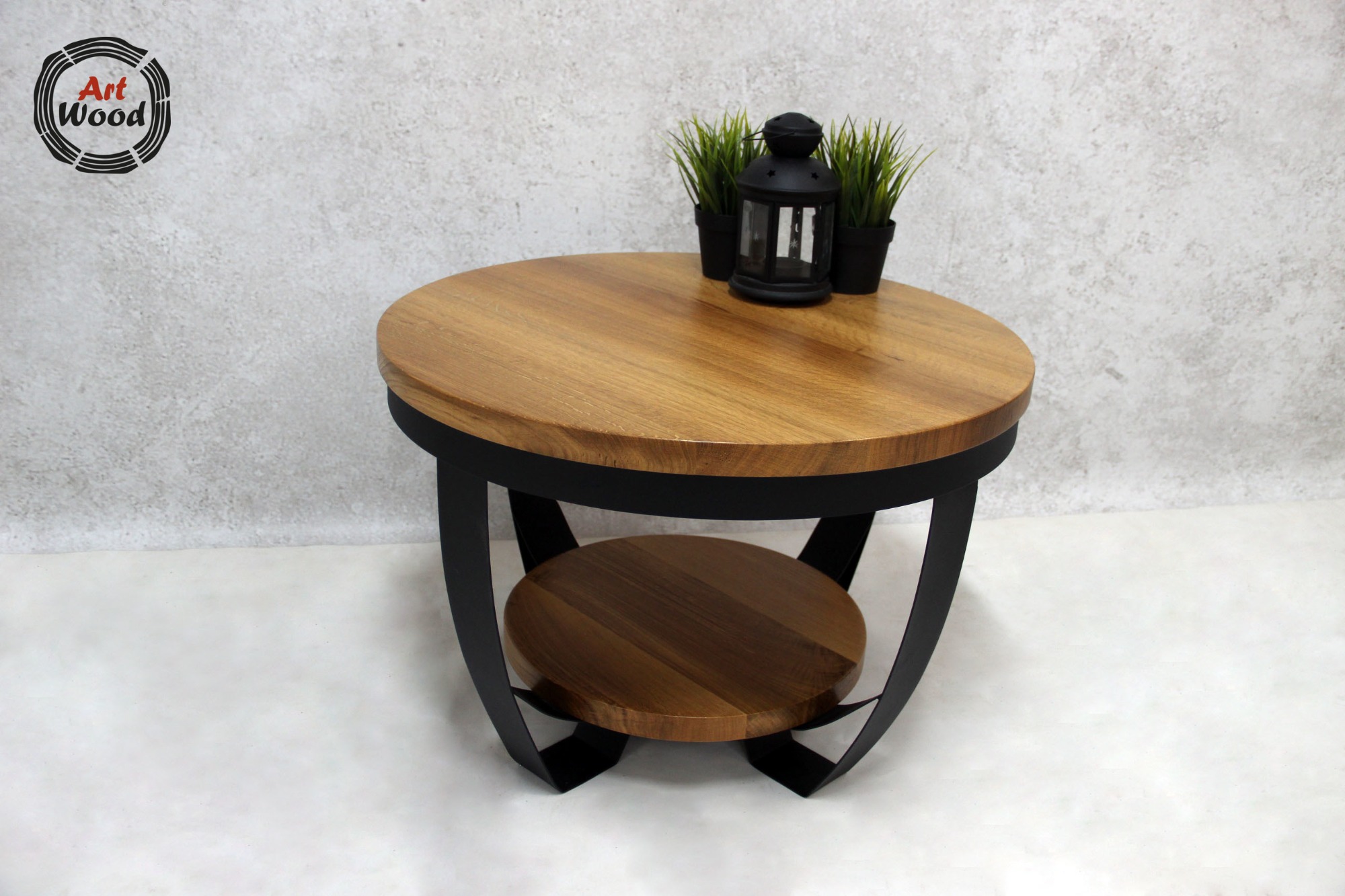 Okrągły stolik kawowy z blatem z jasnego drewna i czarną, metalową podstawą z półką, na blacie lampion i dwie małe doniczki z zielonymi roślinami, w tle szara ściana, w lewym górnym rogu logo firmy...