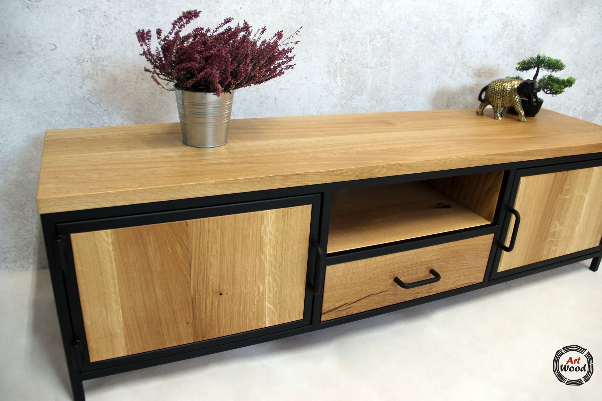 Stolik RTV z drewna dębowego i czarnego metalu, z szufladą i szafkami, dekorowany wrzosem w metalowej doniczce i figurką słonia z bonsai, na tle szarej ściany.
