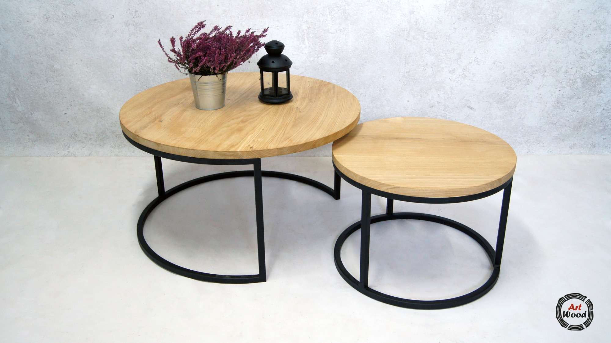 Dwa okrągłe stoliki kawowe z jasnego drewna z czarnymi metalowymi nogami, na większym stoliku dekoracje - wrzos w metalowej osłonce i mała czarna latarenka, w prawym dolnym rogu logo firmy.