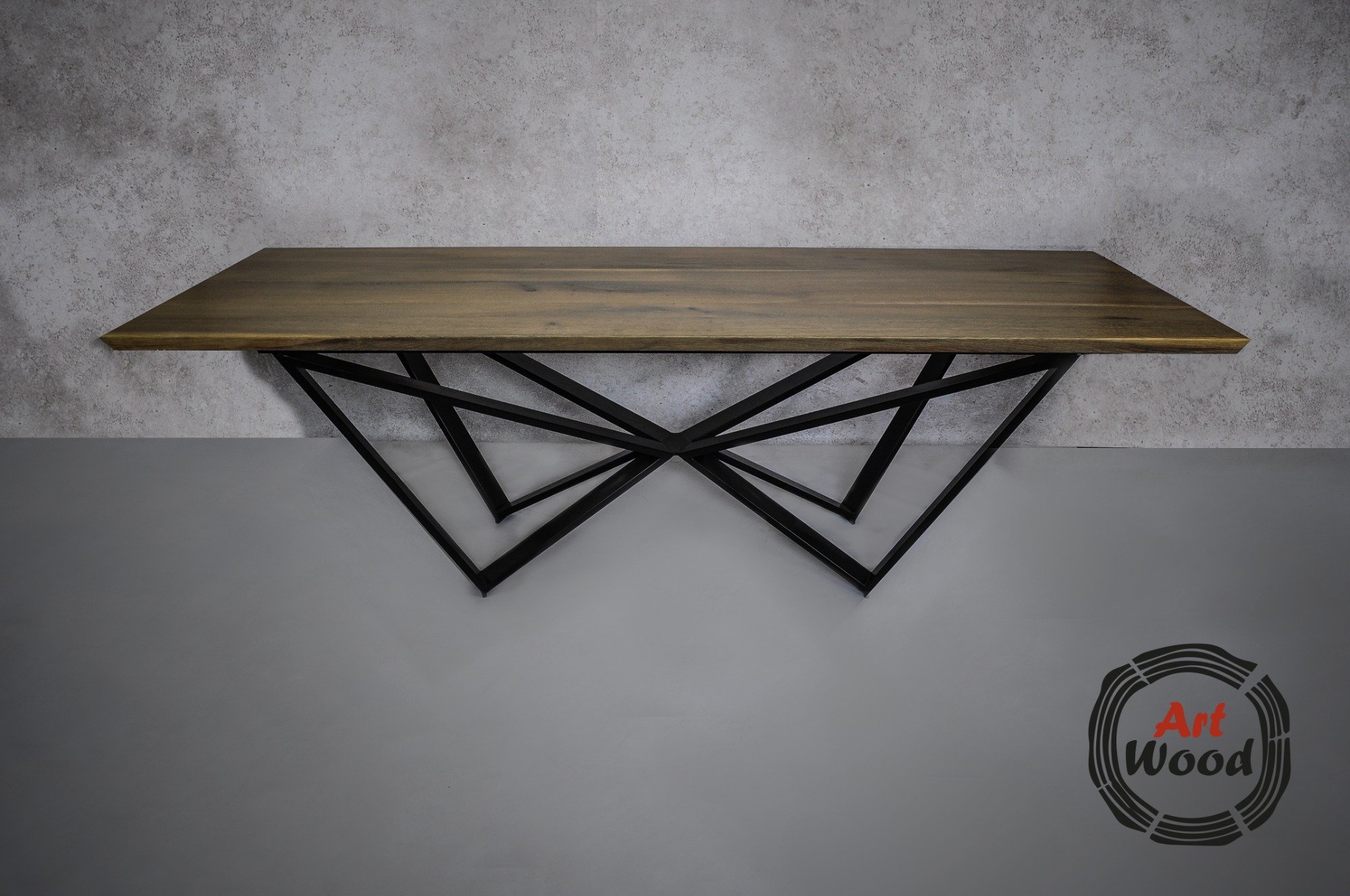 Stół z blatem z ciemnego drewna i geometryczną, metalową podstawą na tle szarej ściany, z widocznym logo firmy w prawym dolnym rogu.
