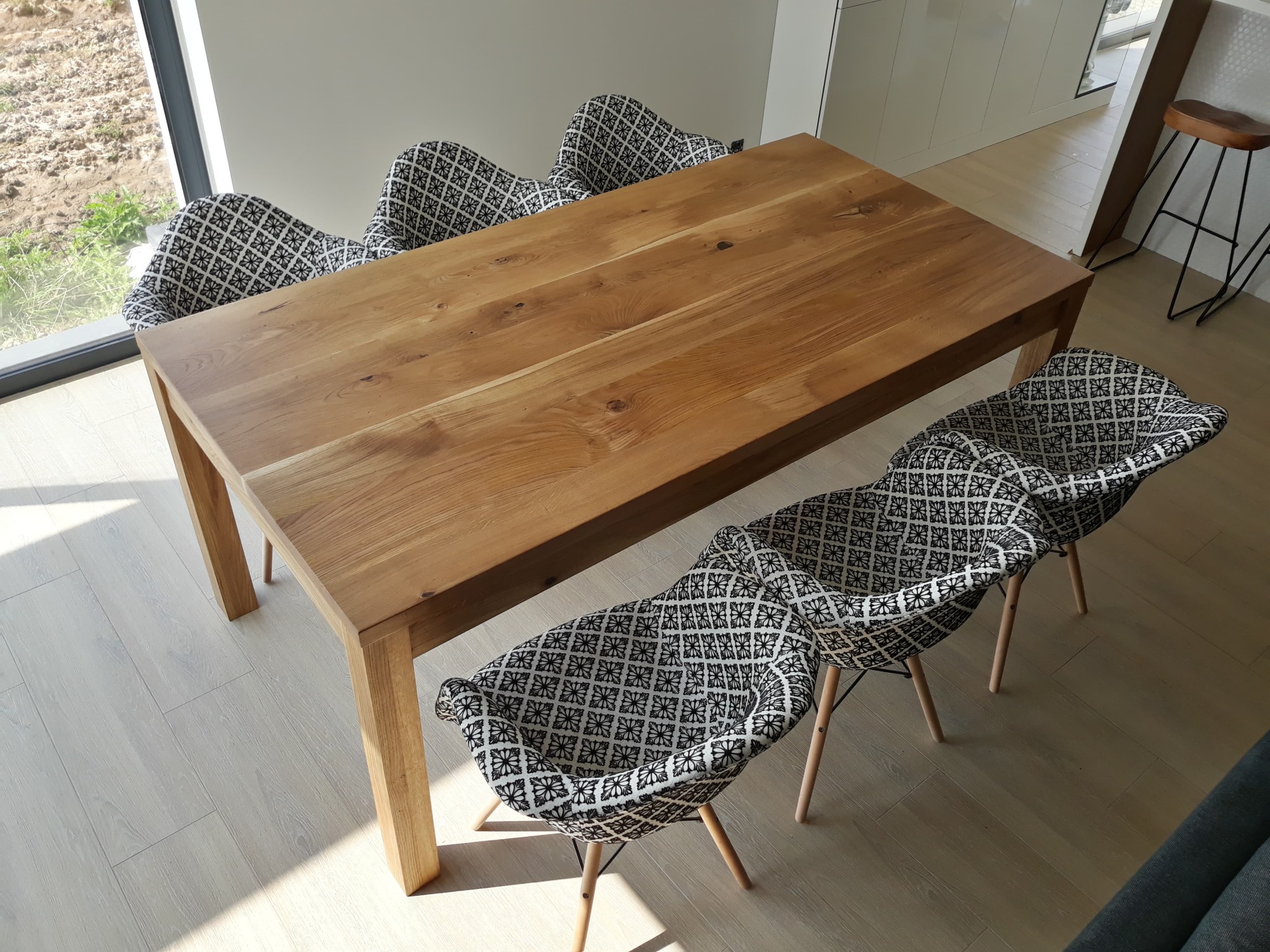 Stół z litego drewna z czterema krzesłami tapicerowanymi w geometryczny wzór, ustawiony w jasnym, nowoczesnym wnętrzu z dużym oknem.