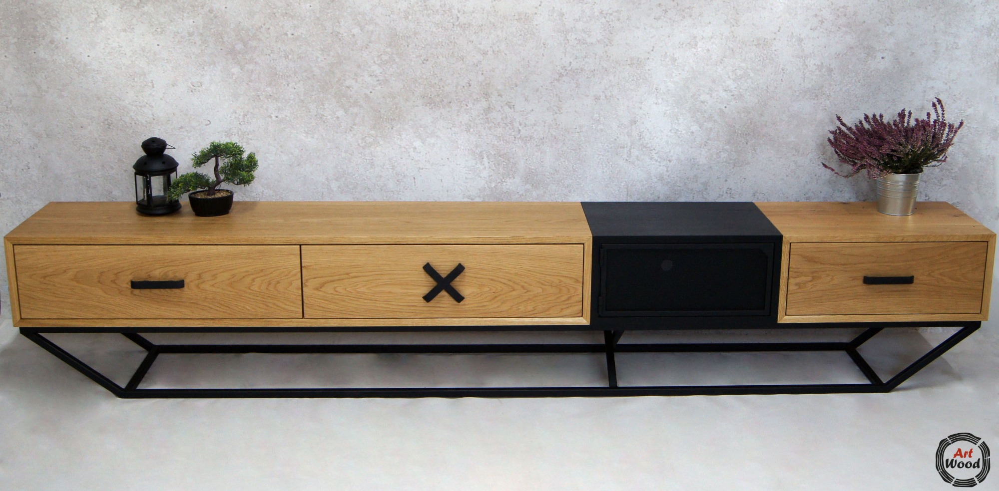 Drewniana szafka RTV z trzema szufladami, jedna czarna, na metalowym stelażu, z dekoracjami: lampion, bonsai i wrzos w metalowej osłonce na blacie, na tle szarej ściany.