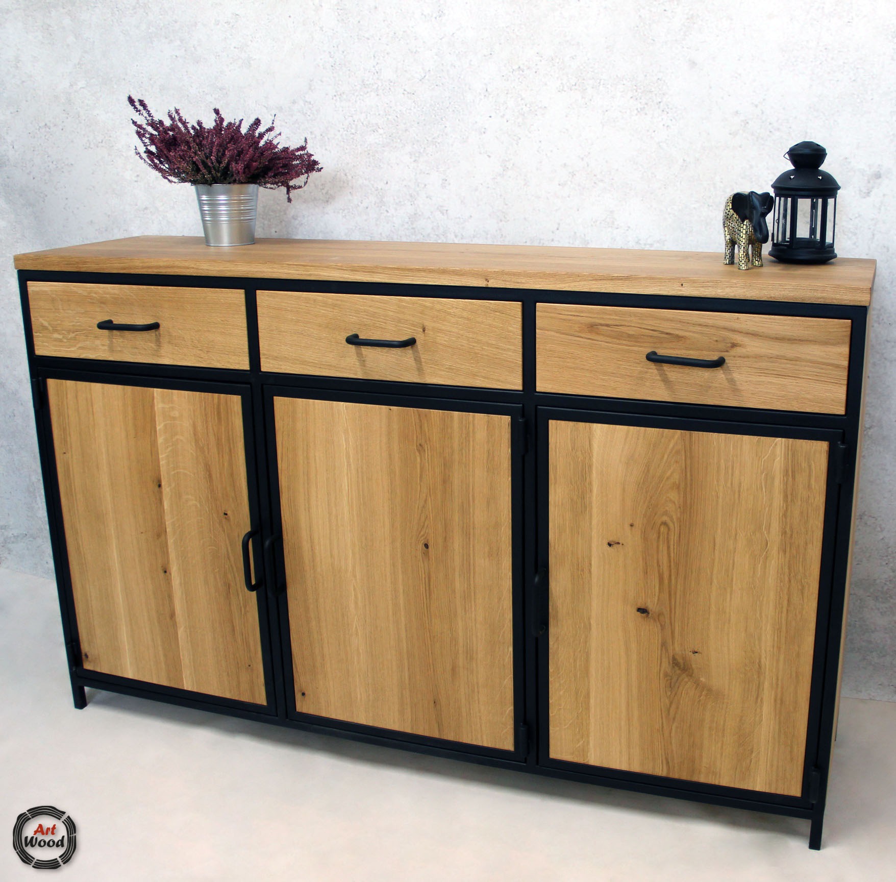 Komoda z trzema szufladami i trzema szafkami z jasnego drewna dębowego, osadzona na czarnej metalowej ramie, z dekoracjami na blacie: bukietem wrzosów w metalowym wiaderku, czarną latarenką...