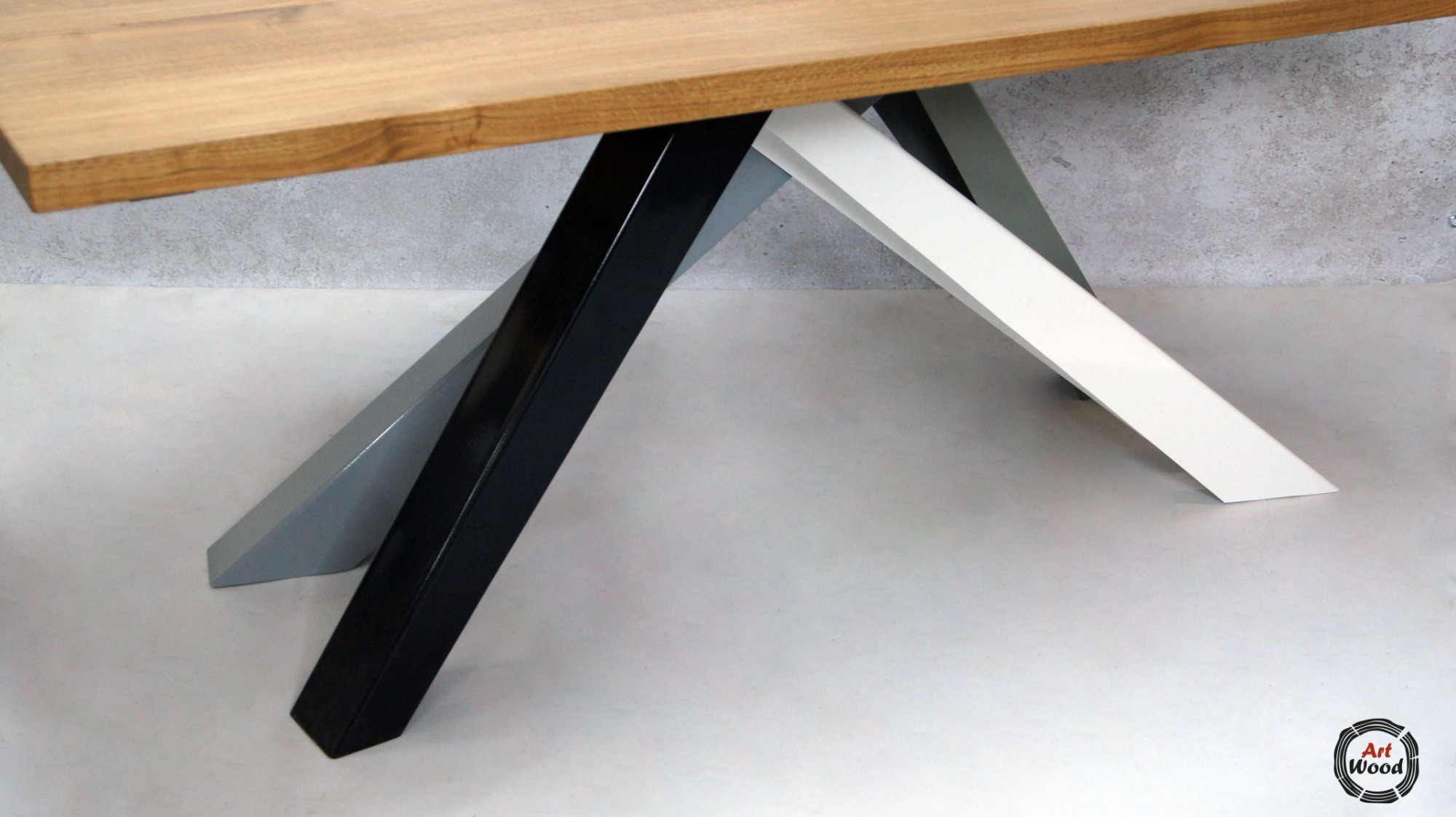 Fragment drewnianego blatu stołu z widocznymi, skośnymi nogami w kolorach: czarnym, białym i szarym, na jasnym tle.