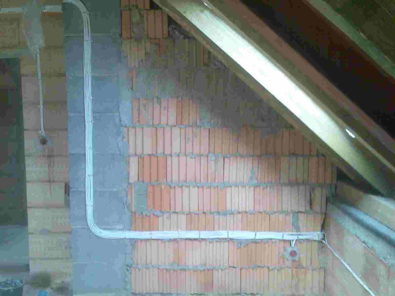 Instalacja elektryczna natynkowa z białymi przewodami ułożonymi w korytkach na surowej ścianie z cegły dziurawki w trakcie remontu poddasza z widoczną konstrukcją drewnianą dachu.