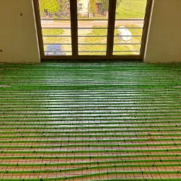 Zielone rury ogrzewania podłogowego ułożone w jodełkę na folii, widok z góry w jasnym pomieszczeniu z oknem. Na zewnątrz zieleń i budynki.