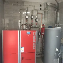 Czerwony piec CO marki Defro z szarym bojlerem i instalacją grzewczą z rur miedzianych i plastikowych na tle szarych płytek ceramicznych.