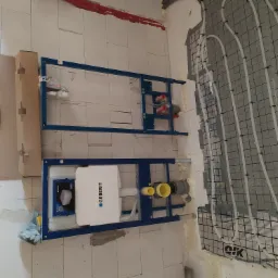 Dwa stelaże podtynkowe Geberit zamontowane na ścianie z białych bloczków, obok widoczne rury ogrzewania podłogowego na siatce, czerwone rury pod sufitem.