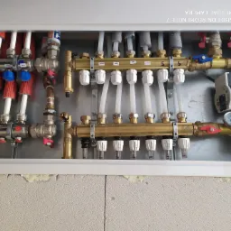 Rozdzielacz ogrzewania podłogowego z widocznymi rurami, zaworami i manometrem zamontowany na szarej ścianie z betonu komórkowego, widoczne napisy 'Łazienka 2' i 'Pokój 2' na naklejkach.