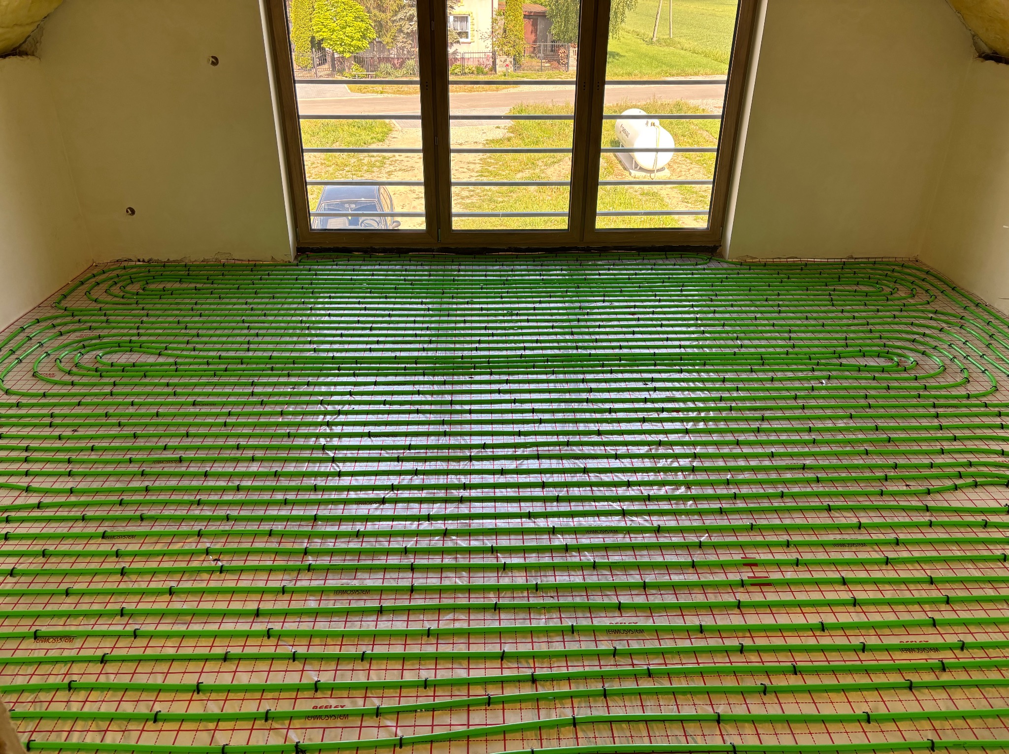 Zielone rury ogrzewania podłogowego ułożone w jodełkę na folii, widok z góry w jasnym pomieszczeniu z oknem. Na zewnątrz zieleń i budynki.