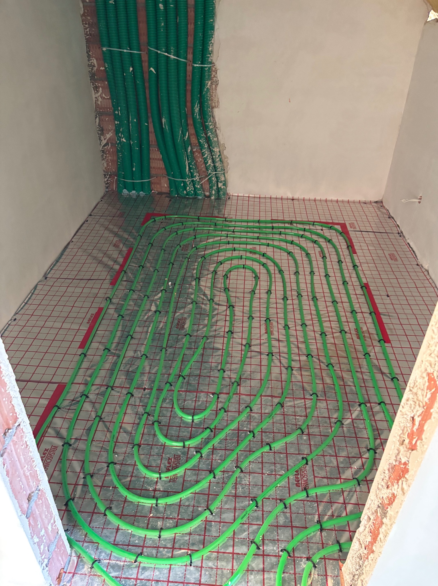 Zielone rury ogrzewania podłogowego ułożone na czerwonej siatce w surowym wnętrzu. Rury pionowe przy ścianie. Widok z góry, bliski kadr.