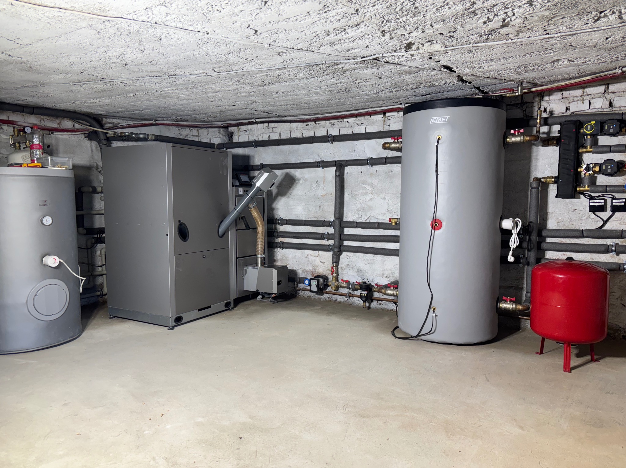 Kompletna kotłownia z szarym piecem, zbiornikiem akumulacyjnym Emei i czerwonym naczyniem przeponowym. Rury w izolacji termicznej biegną wzdłuż ściany z cegły.