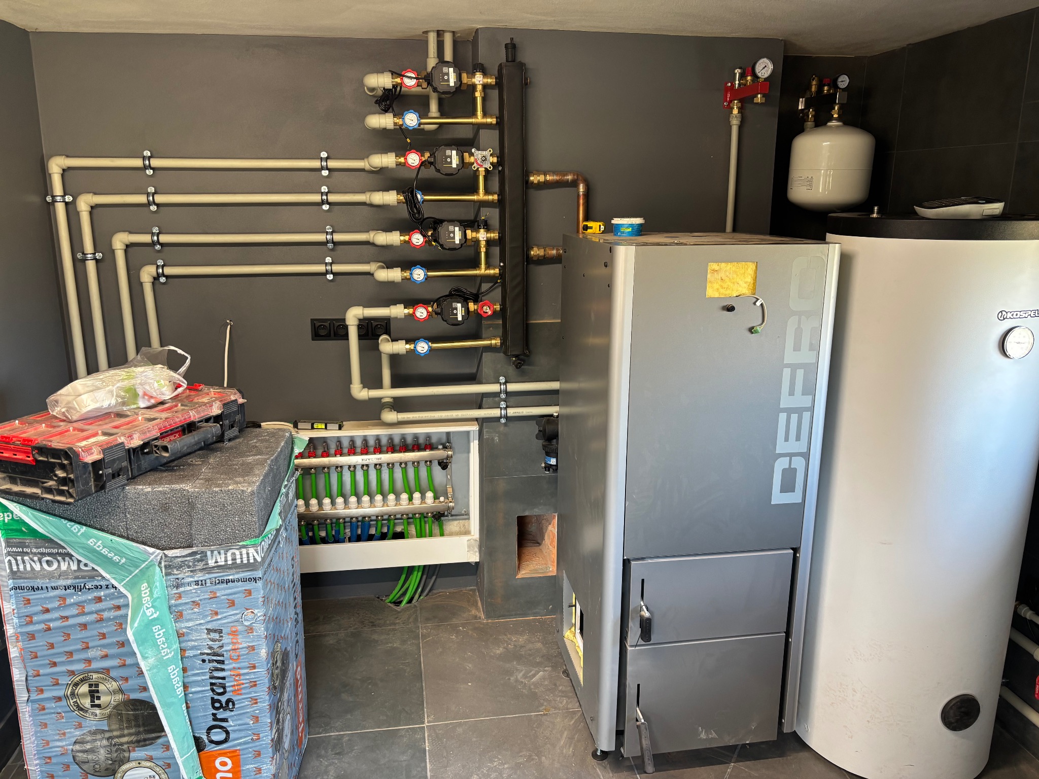 Złożona instalacja grzewcza z piecem Defro i zasobnikiem Kospel, widoczne rury, zawory, manometry i rozdzielacz ogrzewania podłogowego. Elementy instalacji zamontowane na szarej ścianie.