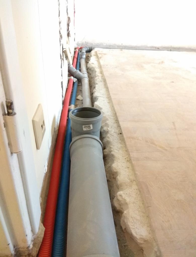 Wykop w podłodze z ułożonymi rurami: szarą kanalizacyjną, niebieską i czerwoną karbowaną. Widoczne przyłącza do ściany i fragment świeżego betonu.