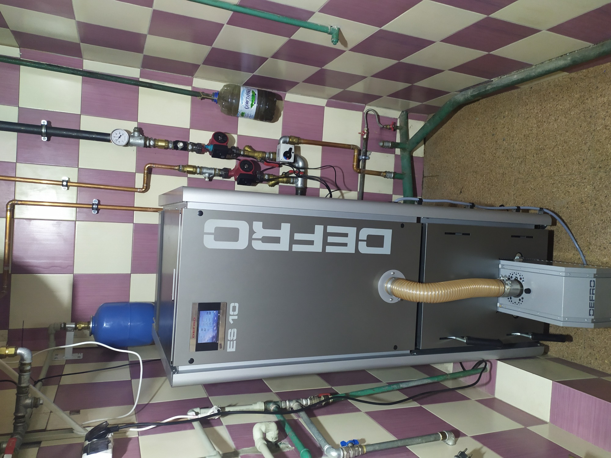 Szary piec grzewczy Defro ES 10 z podłączonymi rurami miedzianymi i plastikowymi, widoczny manometr, butelka z płynem na rurze, tło z płytek w szachownicę.