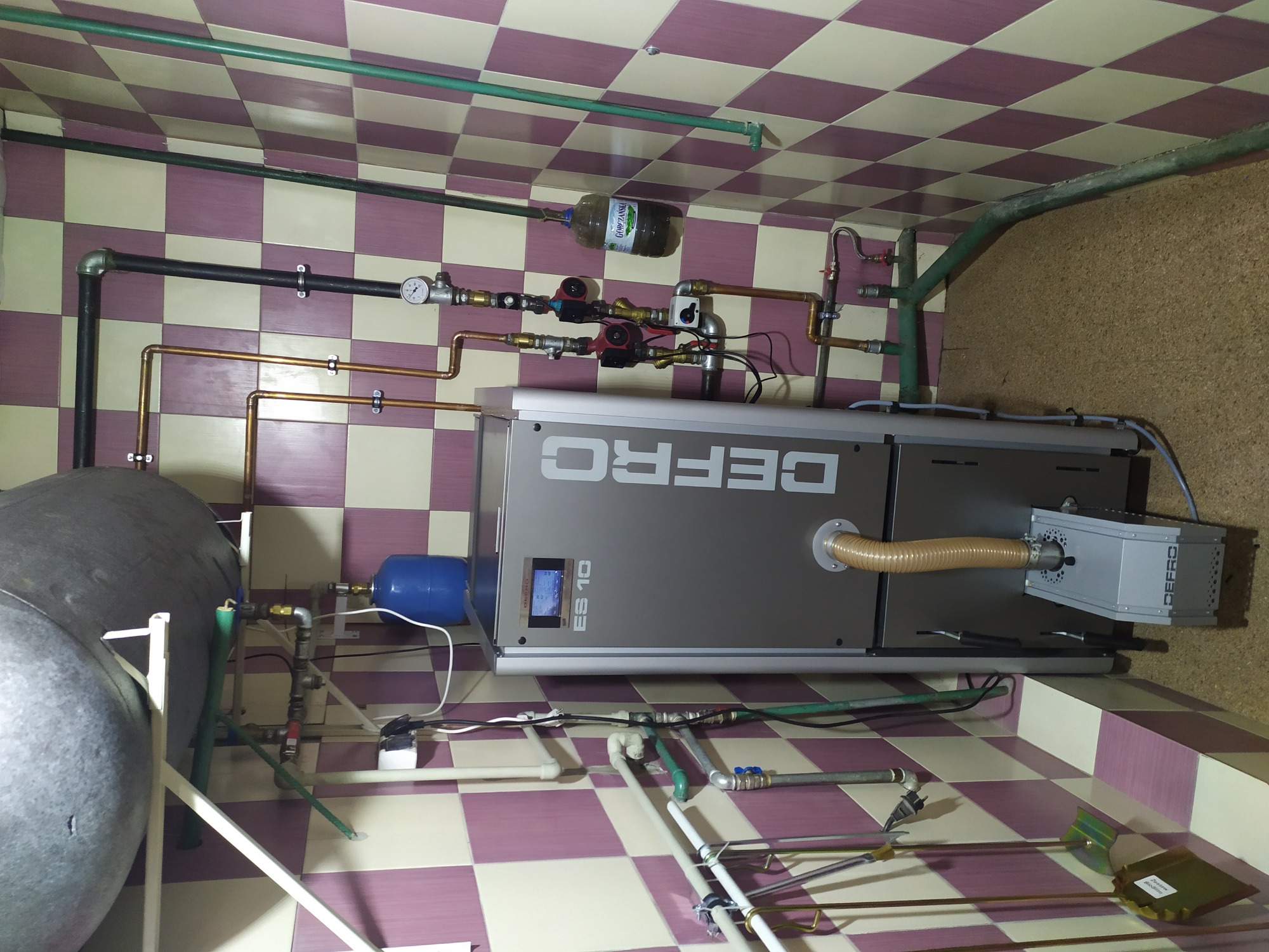 Szary piec DEFRO ES 10 z podłączonymi rurami miedzianymi i zbiornikiem akumulacyjnym w pomieszczeniu z kafelkami w szachownicę.