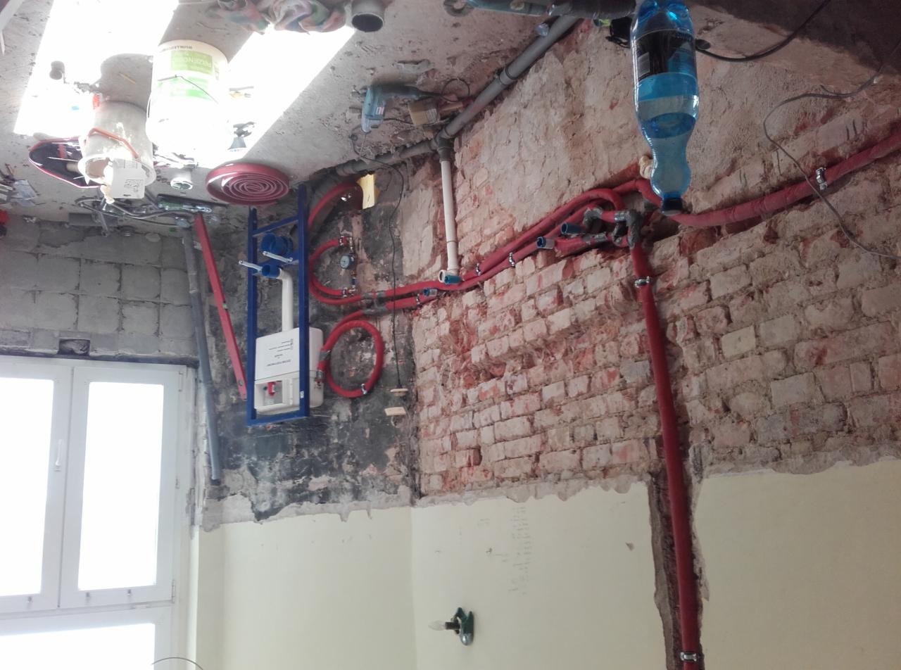 Remontowana łazienka: widok na instalację podtynkową WC, częściowo odsłoniętą cegłę na ścianie, oraz nowe rury instalacyjne w kolorze czerwonym.
