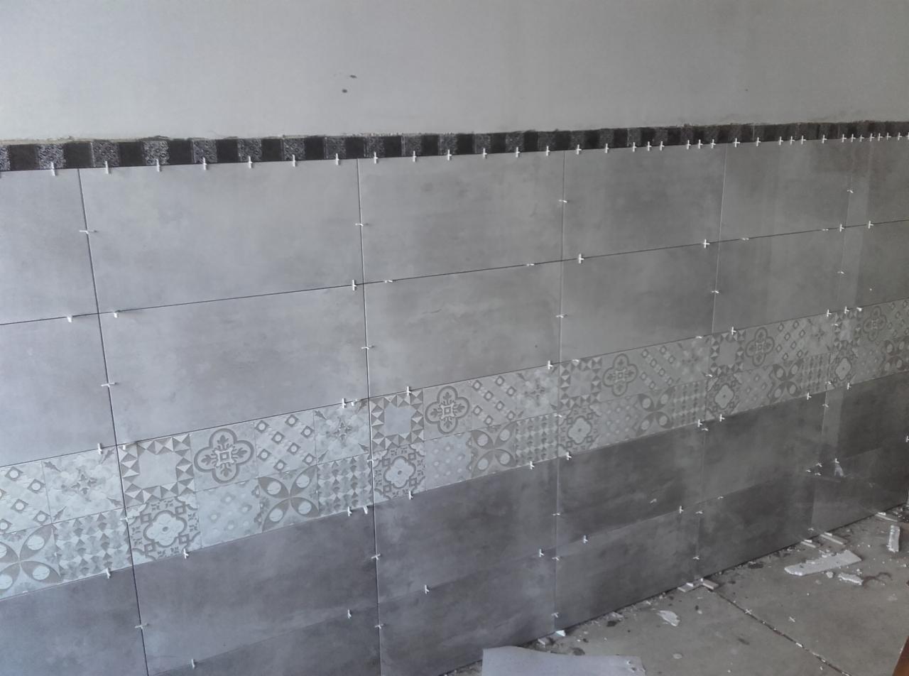 Ściana w trakcie układania szarych płytek z dekoracyjnym pasem wzorzystych płytek, widoczne krzyżyki dystansowe i niedokończona górna krawędź.