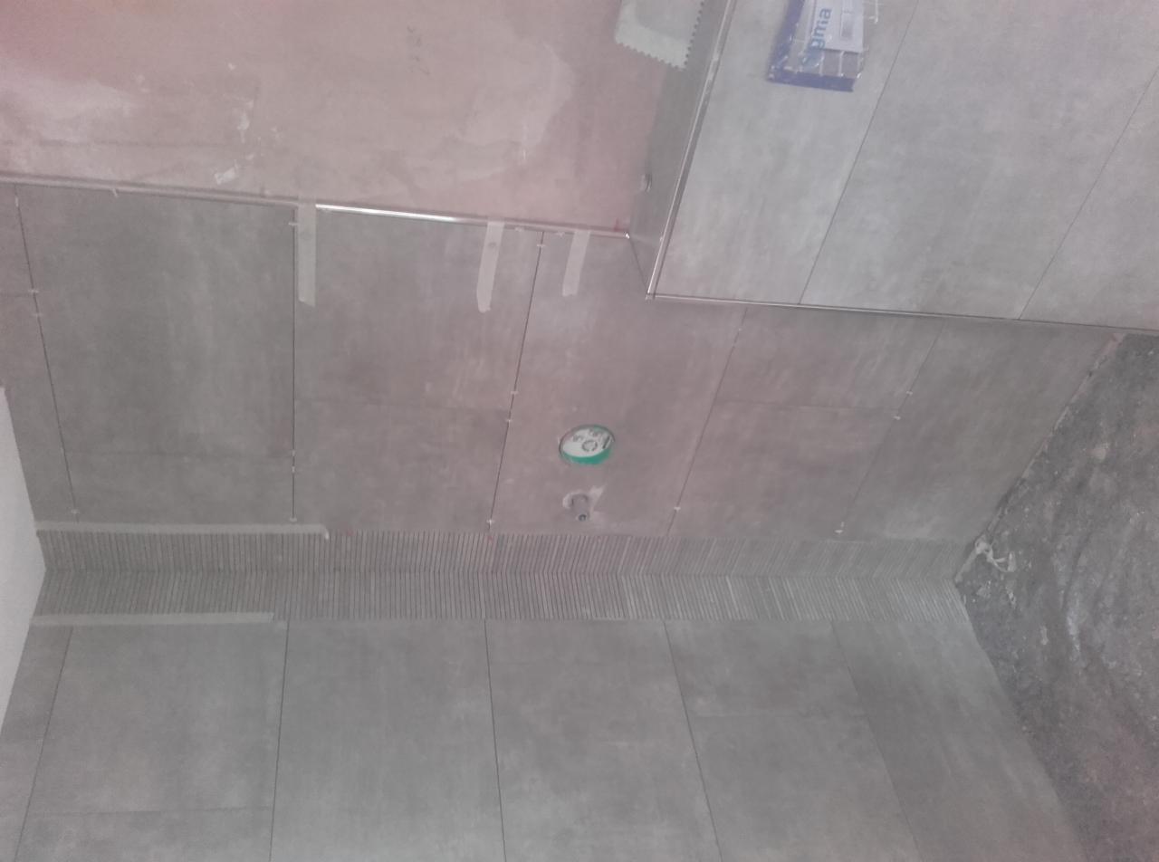 Układanie szarych płytek w łazience, widoczne taśmy mocujące, częściowo wykończona ściana z dekoracyjnym pasem, surowy sufit i przygotowane miejsce na oświetlenie.