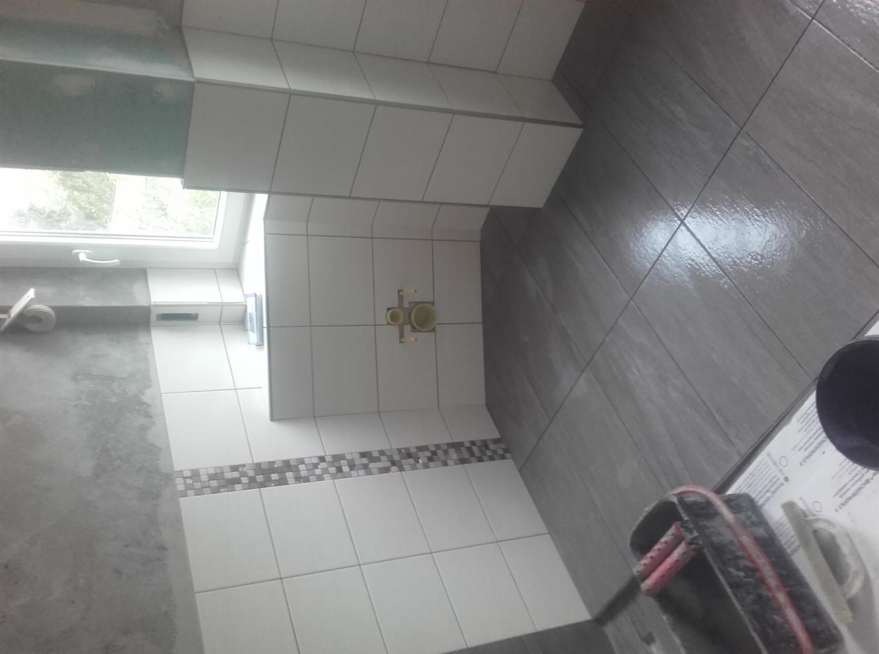 Wnętrze łazienki w trakcie prac wykończeniowych: białe płytki ścienne z dekoracyjnym pasem mozaiki i szare płytki podłogowe, widoczne elementy instalacji wodnej oraz narzędzia glazurnicze.