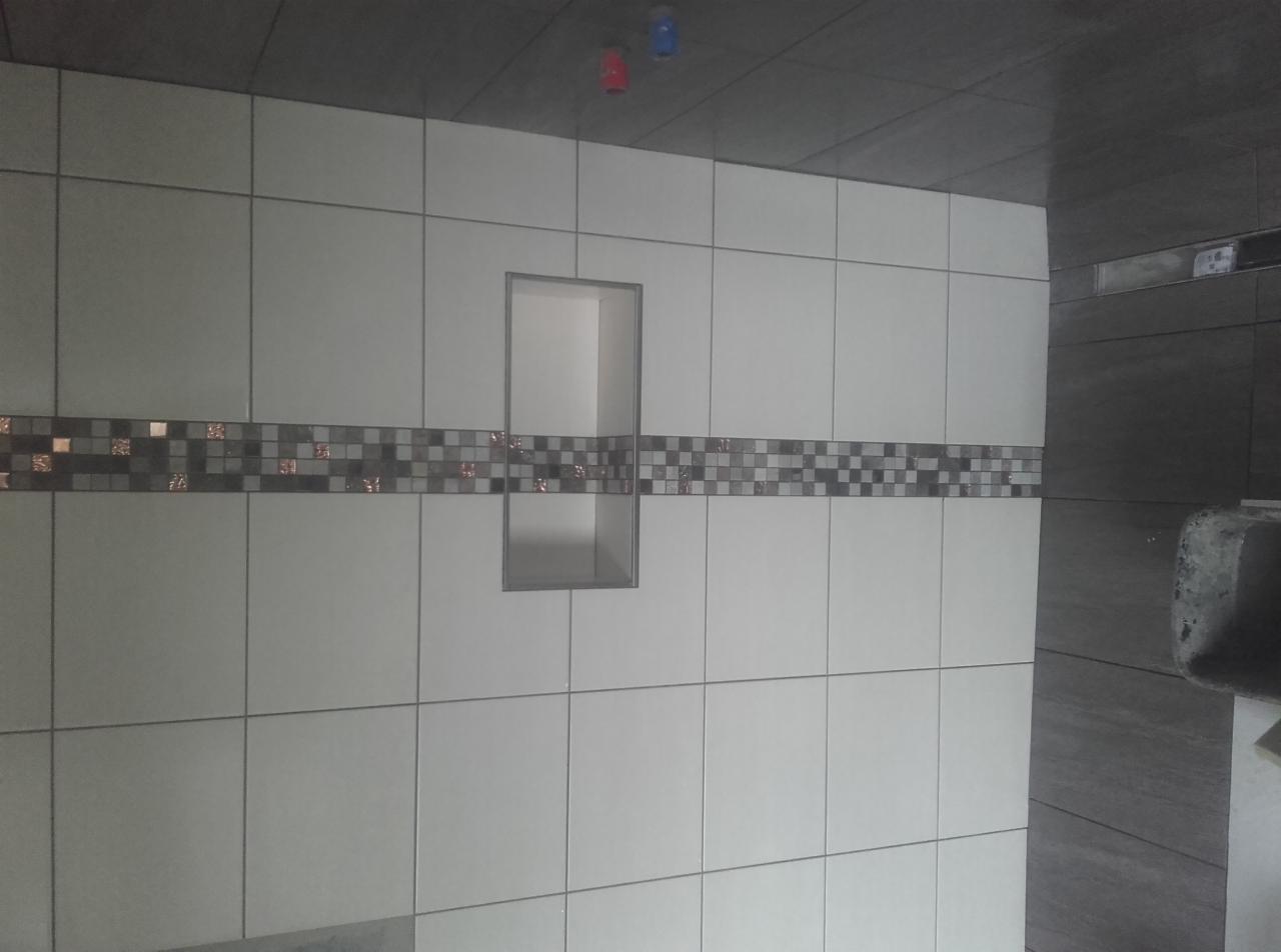 Wykończona ściana z białych płytek w łazience z poziomym pasem dekoracyjnej mozaiki i wnęką w ścianie.