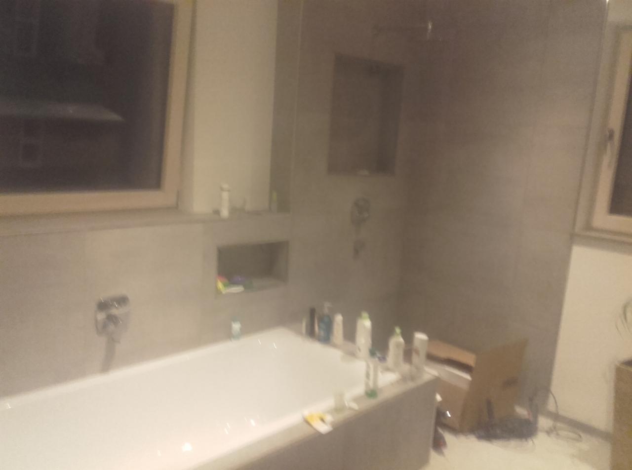 Nowoczesna, jasna łazienka w Szczecinie z wanną, szarą glazurą na ścianach, wnękami na kosmetyki i oknem, częściowo zagracona podczas prac wykończeniowych.