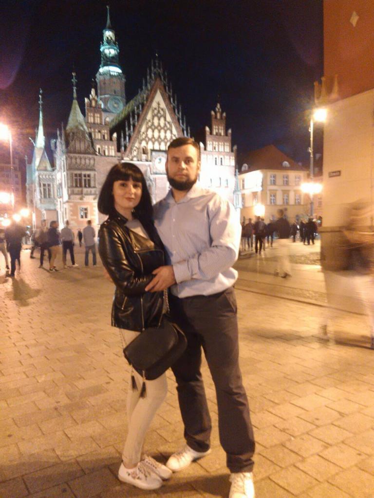 Para pozuje na tle oświetlonego Ratusza we Wrocławiu nocą. Kobieta w skórzanej kurtce i jasnych spodniach, mężczyzna w koszuli i ciemnych spodniach, oboje w białych butach sportowych.