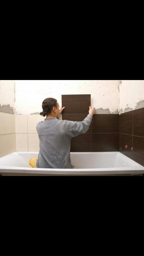 Hydraulik w szarej bluzie, stojąc w wannie, układa brązowe płytki na ścianie łazienki podczas remontu.