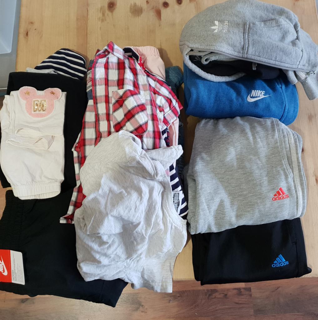 Stos ubrań dziecięcych i młodzieżowych na drewnianym stole, widoczne marki Adidas i Nike, różnorodność kolorów i wzorów, odzież złożona w schludne kupki.