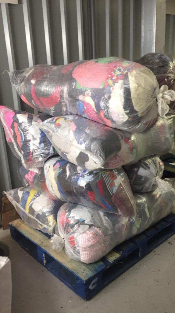 Stos worków z sortowaną odzieżą, zapakowanych w przezroczyste worki foliowe i ułożonych na niebieskiej, drewnianej palecie w magazynie z blachy falistej.