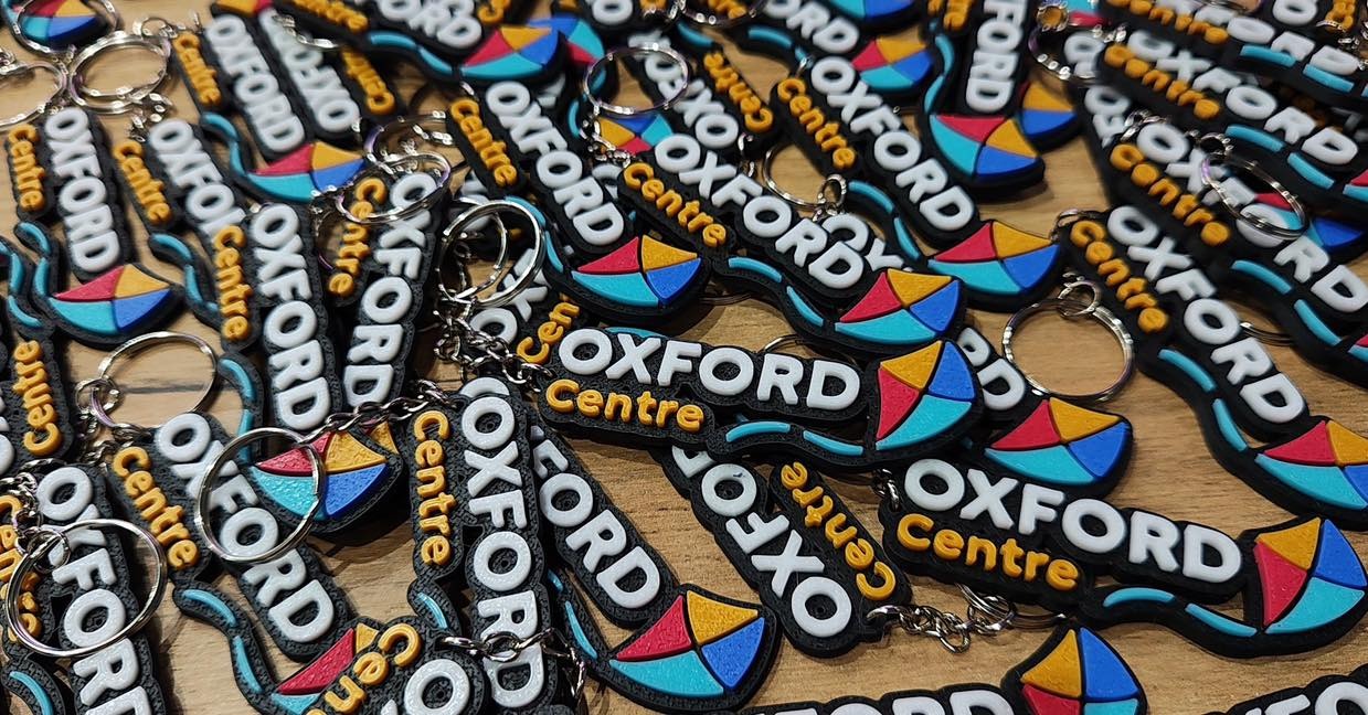 Guma do kluczy z napisem Oxford Centre, czarne tło, białe litery, kolorowy motyw graficzny, ułożone w stos na drewnianym blacie.