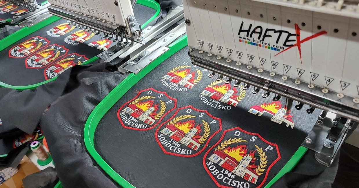 Wielogłowicowa maszyna do haftowania w trakcie wykonywania naszywek z logo OSP Sobocisko na czarnej tkaninie, widoczne tamborki i igły maszyny.