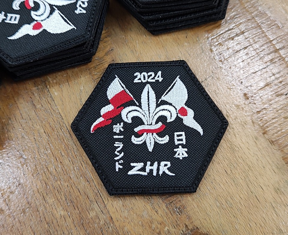 Czarna, sześciokątna naszywka ZHR z wyhaftowanymi flagami Polski i Japonii, lilią harcerską oraz napisem '2024' i nazwami krajów w językach polskim i japońskim, leżąca na drewnianym stole.