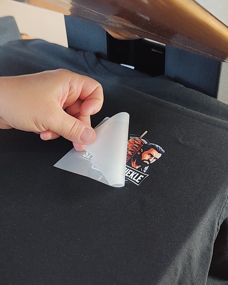 Naklejanie grafiki z motywem brodatego mężczyzny i napisem 'WEKLE' na czarną koszulkę za pomocą termoprasy, usuwanie folii transferowej.