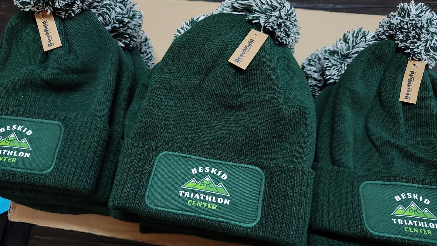Trzy zielone, dziergane czapki zimowe z białymi pomponami i naszywkami 'Beskid Triathlon Center' oraz metkami 'Beechfield Original Headwear'.