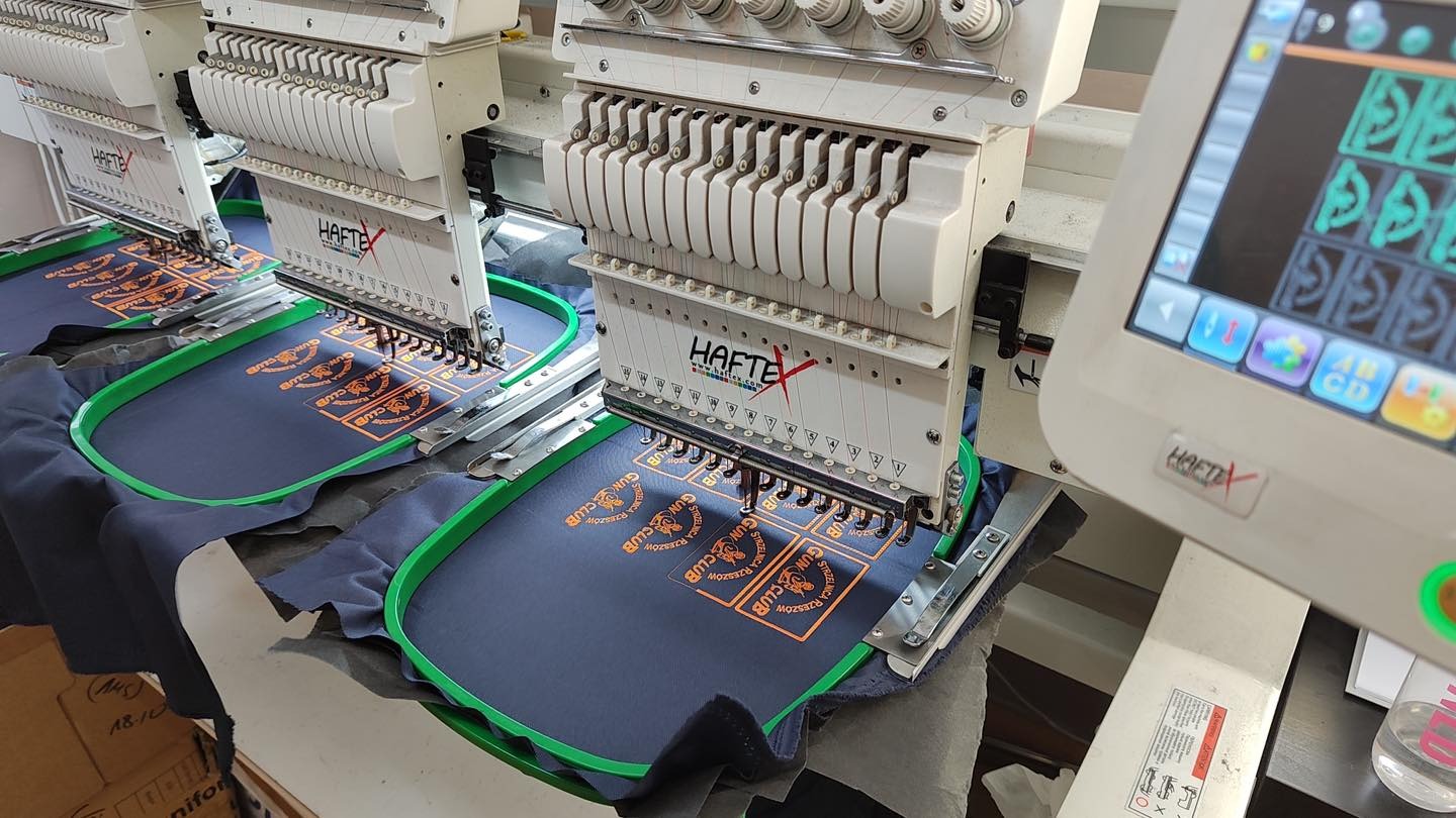 Automatyczna maszyna do haftowania z pomarańczowym wzorem 'GUN CLUB RZESZOW' na ciemnoniebieskiej tkaninie, widoczne igły w trakcie pracy i interfejs maszyny.