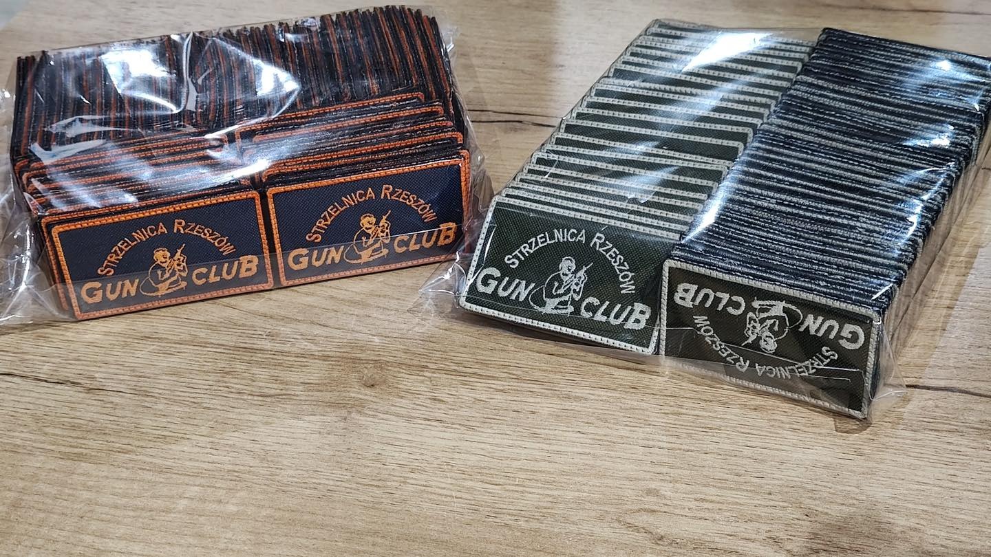 Dwa stosy naszywek z napisem 'Strzelnica Rzeszów Gun Club' w różnych kolorach, zapakowane w folię, leżące na drewnianym blacie.
