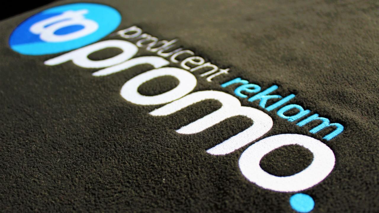 Ciemny materiał z wyhaftowanym białym napisem 'promo' i niebieskim napisem 'producent reklam', widoczne zbliżenie na teksturę materiału i precyzję wykonania haftu.