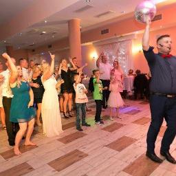 Sweet Times - Wodzirej z mikrofonem i czerwoną muchą trzymający dyskotekową kulę nad głową, prowadzi zabawę taneczną dla grupy gości w sali bankietowej z parkietem imitującym drewno.