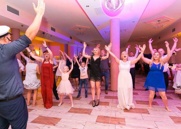 Rozbawiona grupa gości weselnych tańczy z podniesionymi rękami na parkiecie, oświetlona dyskotekową kulą i kolorowymi światłami, z widocznym mężczyzną w kapitańskim stroju z przodu.
