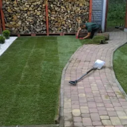 Układanie trawy z rolki w ogrodzie: widoczny fragment ułożonej trawy, pracownik w trakcie pracy, stos drewna w tle, ścieżka z kostki brukowej i szpadel leżący na ścieżce.