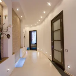 Nowoczesny korytarz z oświetleniem punktowym w podłodze i suficie, dekoracyjną ścianą z teksturą, wazą z kwiatami w niszy oraz drewnianymi drzwiami z matowym szkłem.