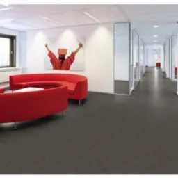 Biuro z ciemną, gładką wykładziną, czerwonymi sofami i szklanymi ściankami działowymi. Oświetlenie sufitowe. Nowoczesny design wnętrza.