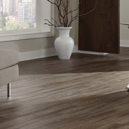 WORK SPOT SPÓŁKA Z OGRANICZONĄ ODPOWIEDZIALNOŚCIĄ - Ciemne, drewnopodobne panele ułożone w salonie z szarą sofą, dekoracyjnym stolikiem i szklanym biurkiem na metalowych nogach. Widok na miasto za oknem.