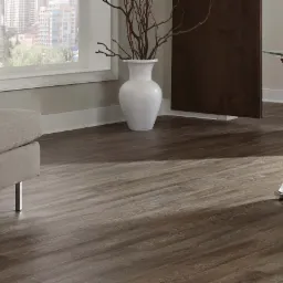 Ciemne, drewnopodobne panele ułożone w salonie z szarą sofą, dekoracyjnym stolikiem i szklanym biurkiem na metalowych nogach. Widok na miasto za oknem.