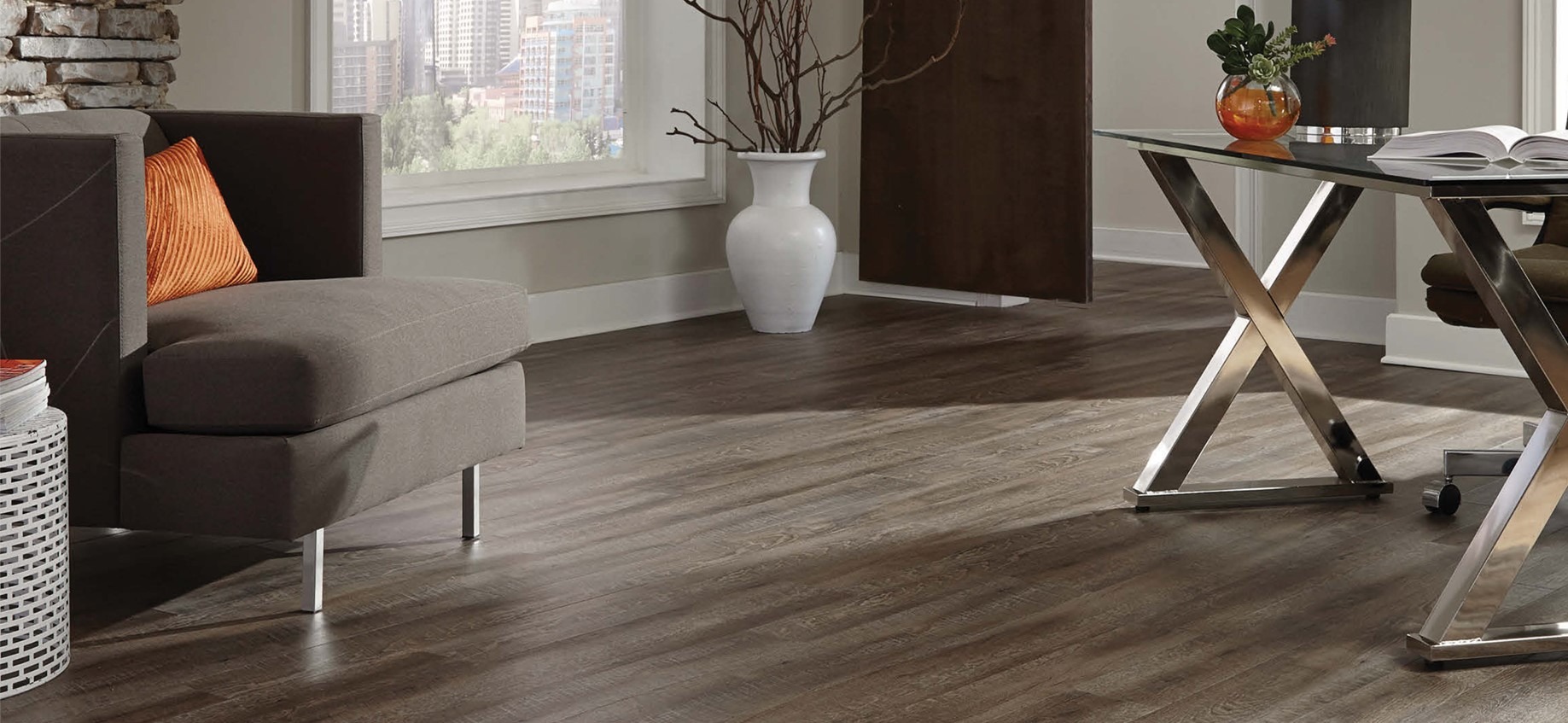 Ciemne, drewnopodobne panele ułożone w salonie z szarą sofą, dekoracyjnym stolikiem i szklanym biurkiem na metalowych nogach. Widok na miasto za oknem.